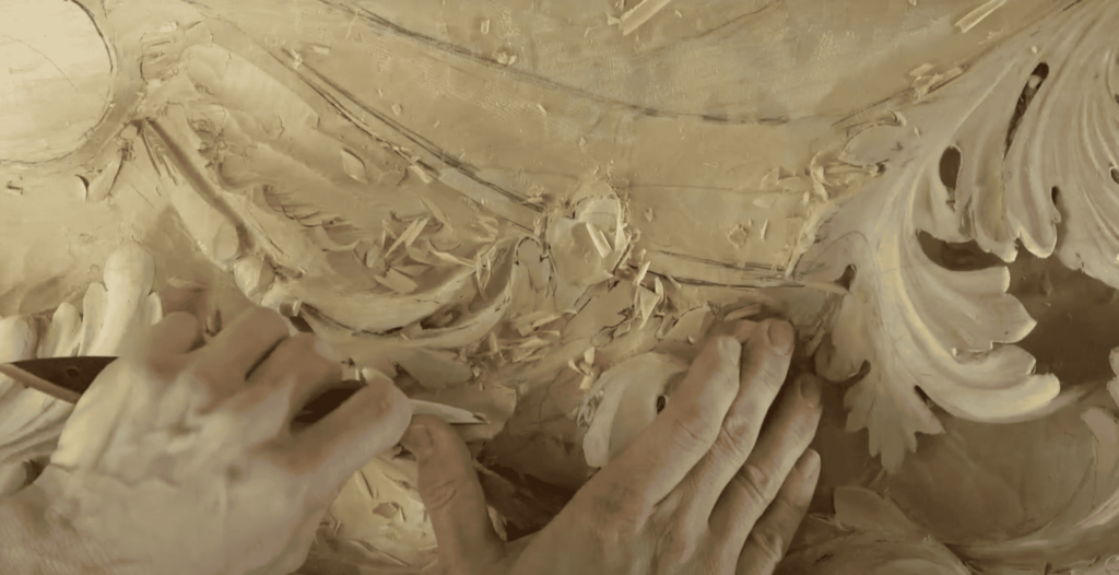 Alt text: Close-up of a person's hands skillfully carving intricate patterns into wood. The focus is on the carving workbench with shavings scattered around, showcasing the detail of the wood carving process. This image captures the essence of wood carving classes and lessons.